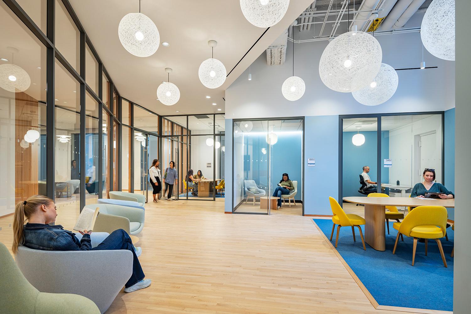 A large windowed office suite is for financial well-being. Students and staff are sitting at tables and lounge chairs working.